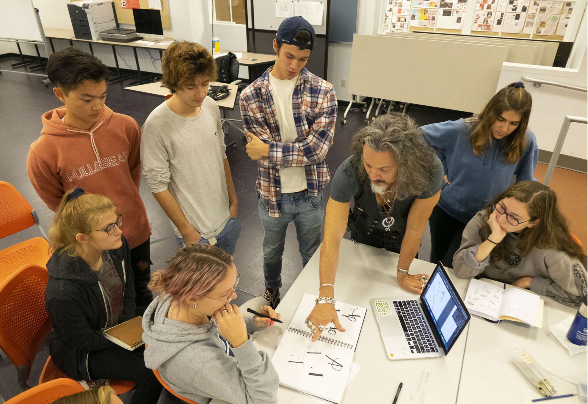 Students gathered around a table with sketches and a laptop open as the instructor points at concepts on the pages