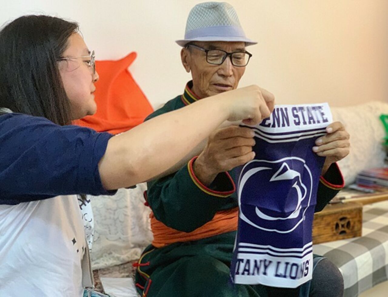 A music education Ph.D. student shows a blue and white Penn State Nittany Lions garden flag to an older individual sitting on a couch.