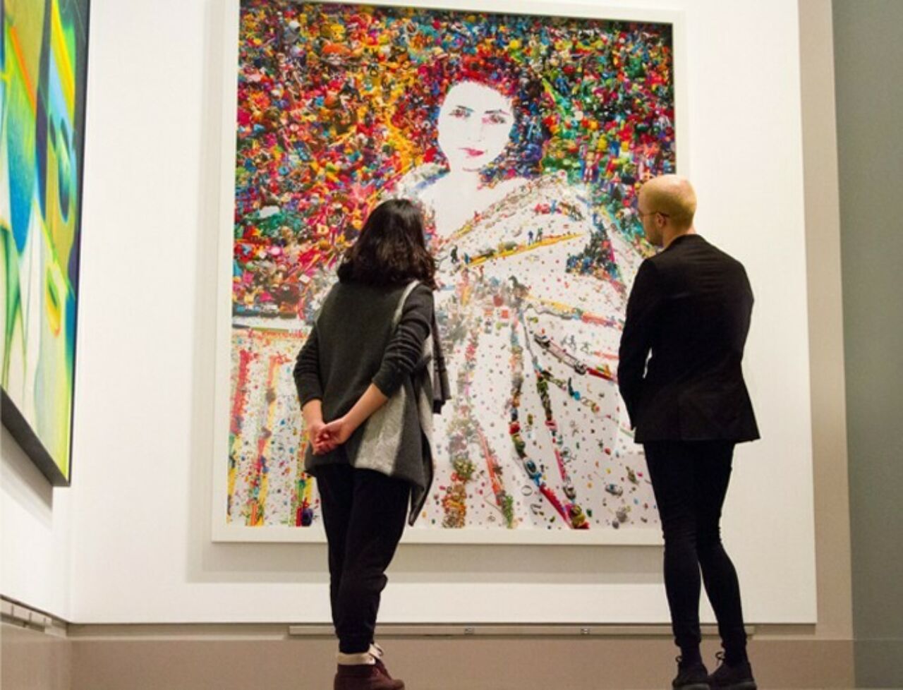 Two people standing in front of a large portrait of a woman with bright colorful pops of color and texture creating the backdrop of the piece, located in the Palmer Museum of Art.