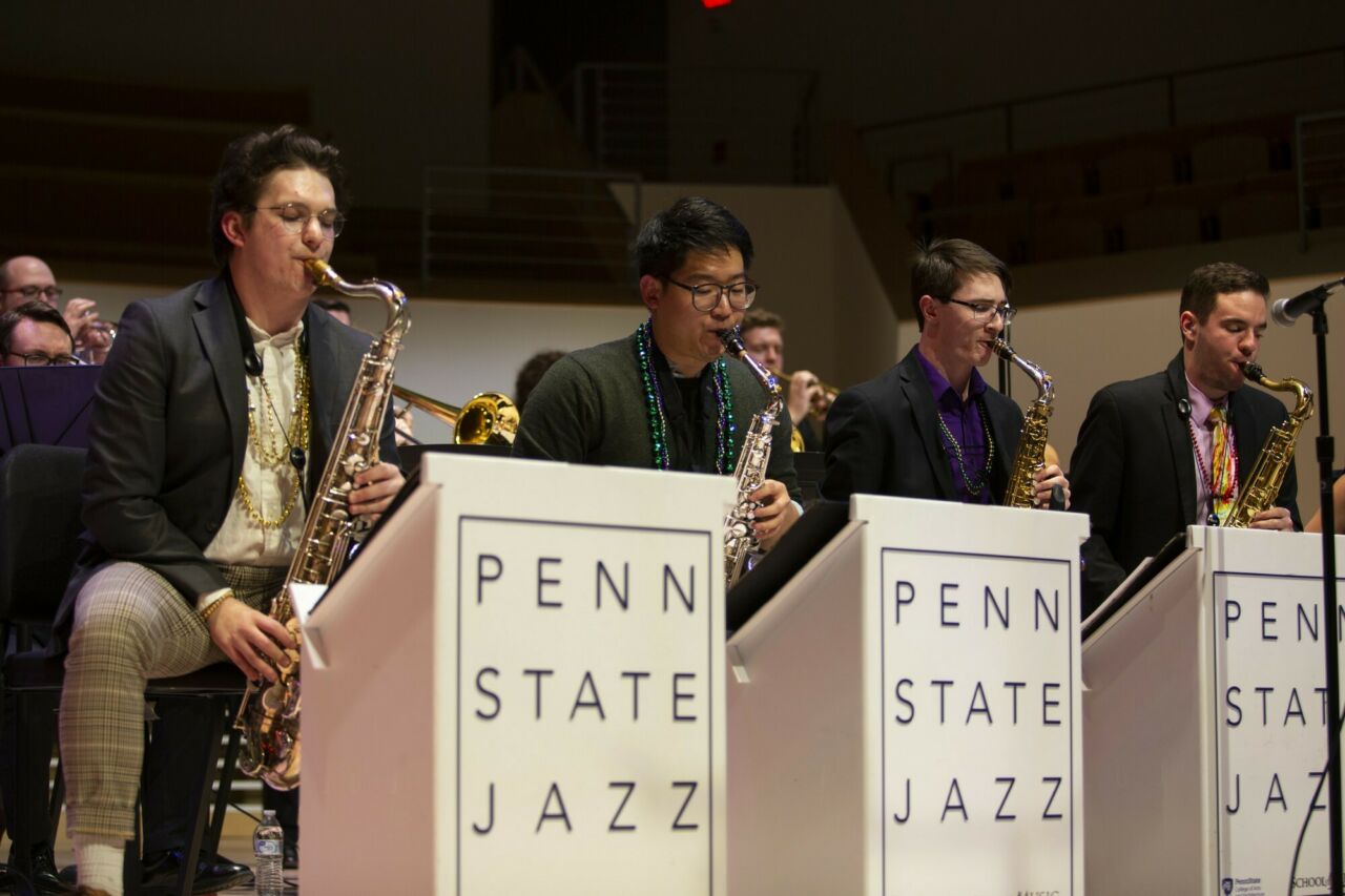 Group of four School of Music students playing the saxophone during a jazz performance, behind a row of Penn State Jazz podiums.