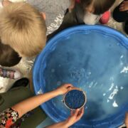 Overhead view of a group of children surrounding a bright blue tub filled with liquid; one student has arms outstretched, holding a copper bowl of what appears to be intense blue dye.