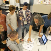 Students gathered around a table with sketches and a laptop open as the instructor points at concepts on the pages