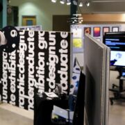 Undergraduate graphic design student work exhibition: showcasing a wall divider with big bold text, digital designs on flat-screen TVs, and poster design pinned up on a divider board.