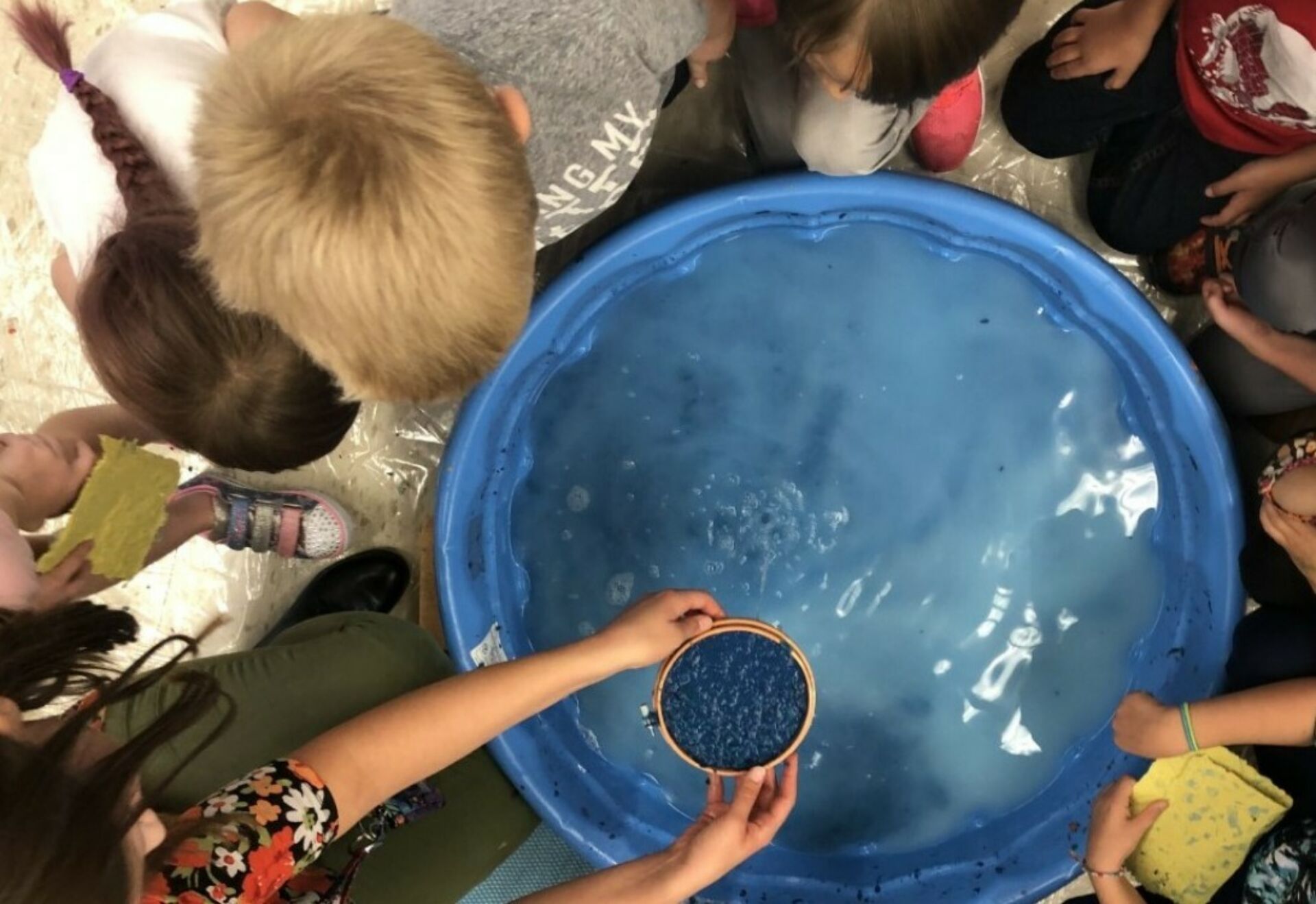 Overhead view of a group of children surrounding a bright blue tub filled with liquid; one student has arms outstretched, holding a copper bowl of what appears to be intense blue dye.