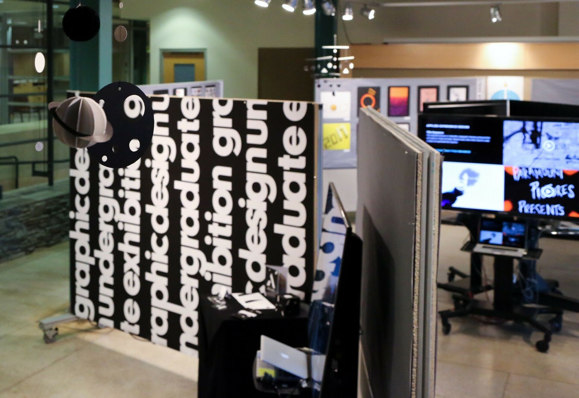 Undergraduate graphic design student work exhibition: showcasing a wall divider with big bold text, digital designs on flat-screen TVs, and poster design pinned up on a divider board.