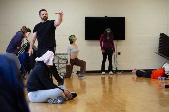 A group of students wearing costume masks posing at various places in a dance studio, their instructor gesturing with an outstretched hand.