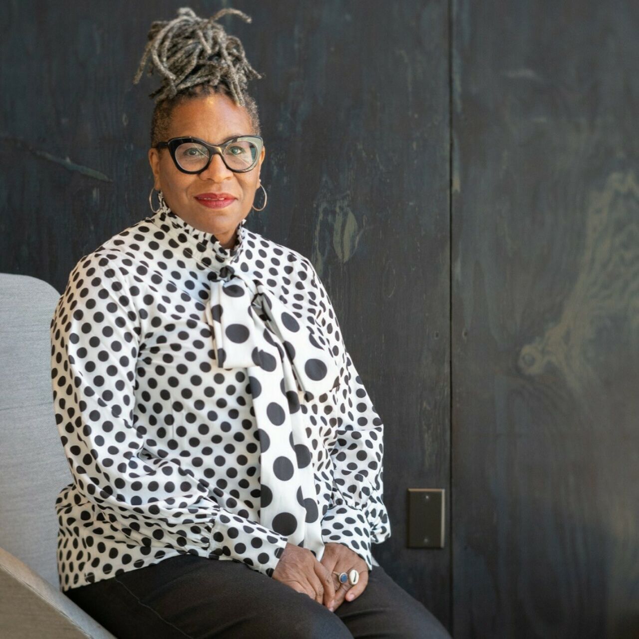 Portrait photo of Folayemi Wilson, a black woman with glasses wearing a white short with black polkadots, sitting in a modern, light-colored chair in front of a dark grey wall.