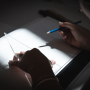 View over the shoulder of a student holding an artist's triangle. and pencil, in process of drawing on a white sheet of paper which is brightly highlighted, with the rest of the image darkened by a significant vignette.