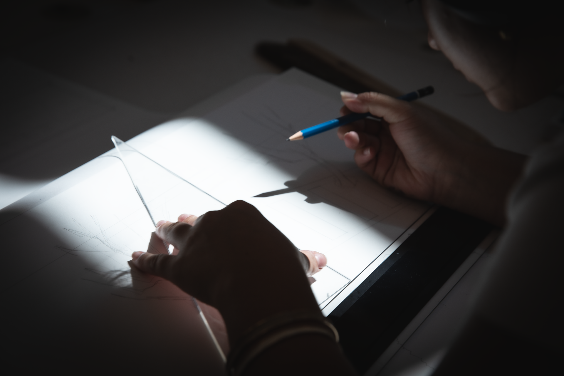 View over the shoulder of a student holding an artist's triangle. and pencil, in process of drawing on a white sheet of paper which is brightly highlighted, with the rest of the image darkened by a significant vignette.