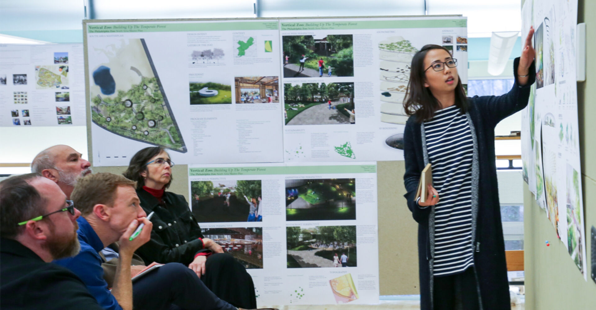 A woman with glasses and dark shoulder-length hair gestures at a board displaying landscape architecture plans. Four members of an audience sit in chairs looking with great focus at the plans as the woman describes them.