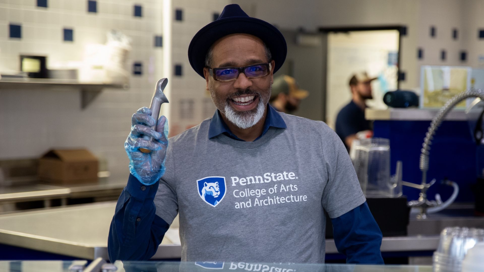 A&A Dean B. Stephen Carpenter II standing behind an ice-cream counter holding a metal scoop aloft and smiling during part of the college's 60th anniversary celebrations.