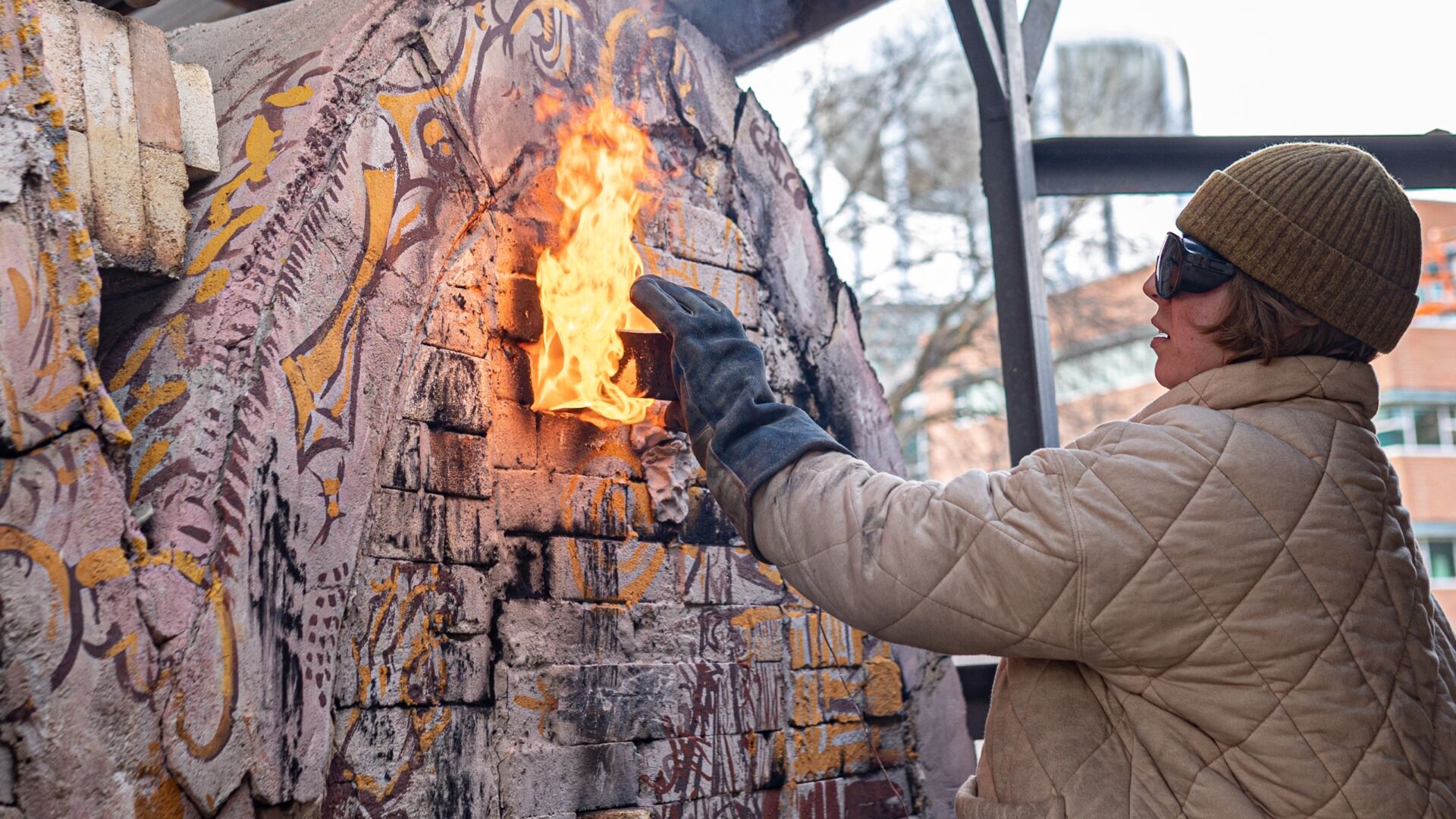 Student wearing gloves, goggles and a heavy coat sliding a brick into a ceramics kiln that has flames rising out of a vent hole.