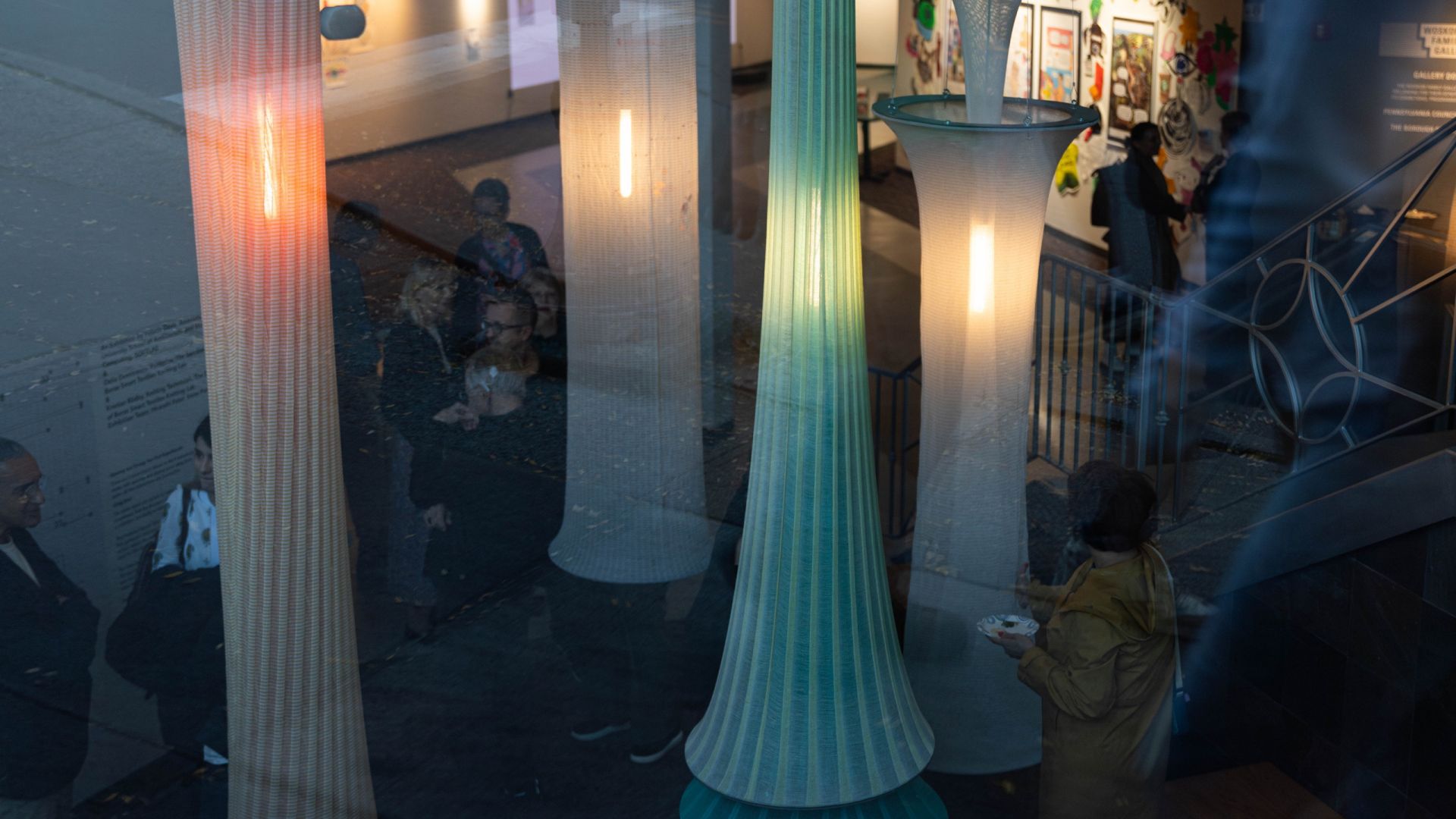 An abstract, overhead view of a gallery space with people gathered around illuminated textile columns. The photo is shot through windows, so there is some complex layering of space and content.