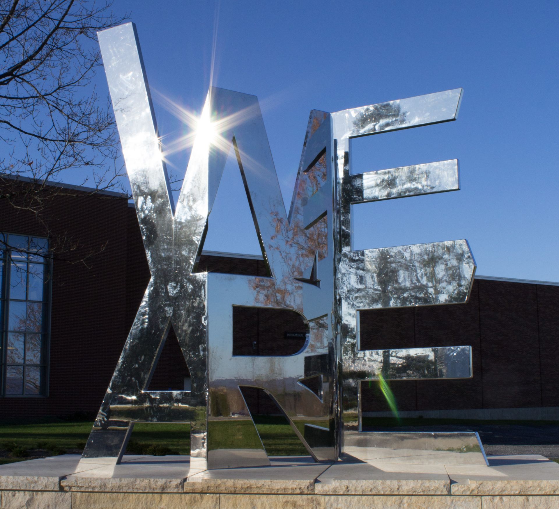 The mirror-finished metallic "We Are" sculpture shining in the sun against a brilliant blue sky background.