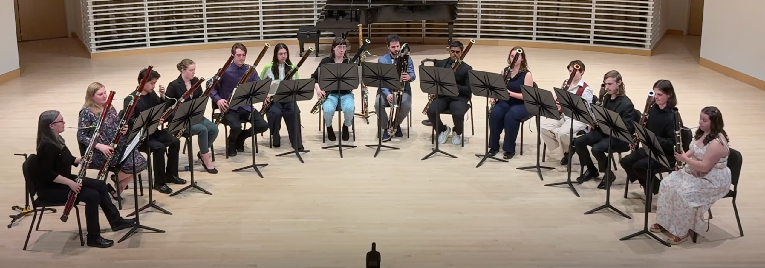 Fourteen bassoonist sit on chairs in a semi-circular arrangement in a recital hall with light natural wooden floor and a grand piano in the background.