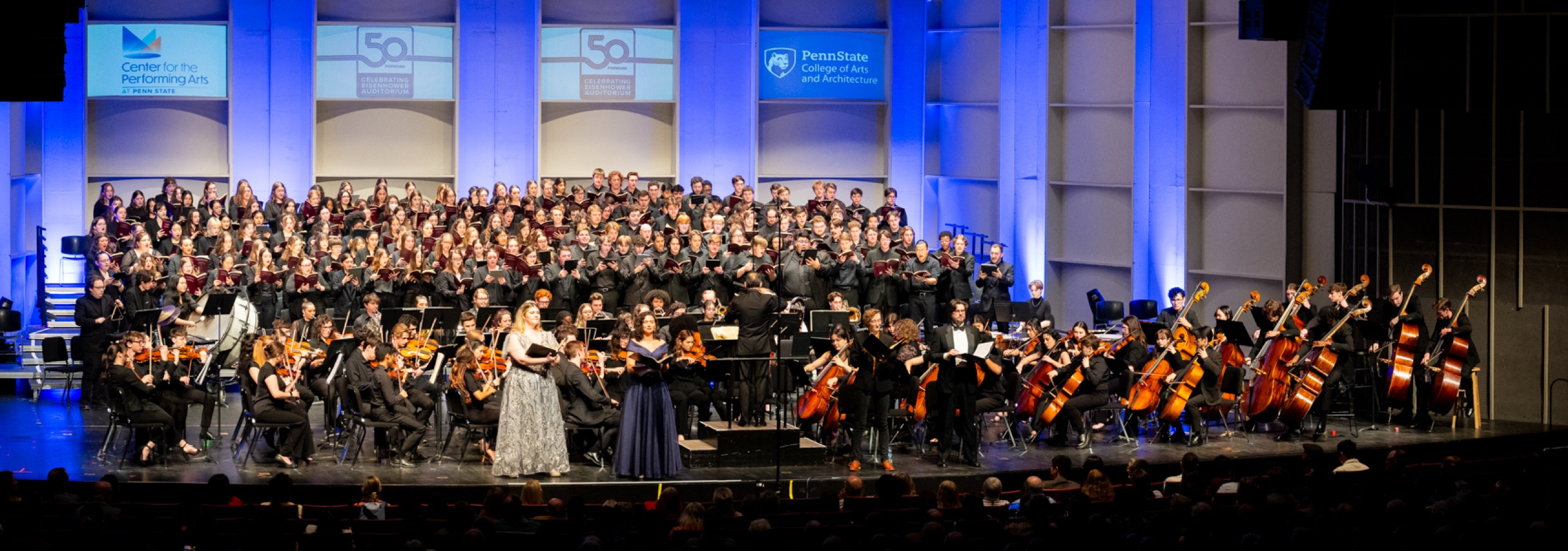 A full orchestra plays on stage in front of a large student choir.