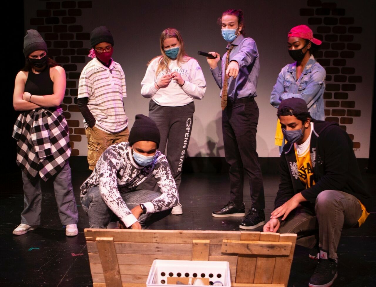 A group of seven diverse student actors huddled around and pointing at a wooden sign, the face of which is not visible to the viewer.
