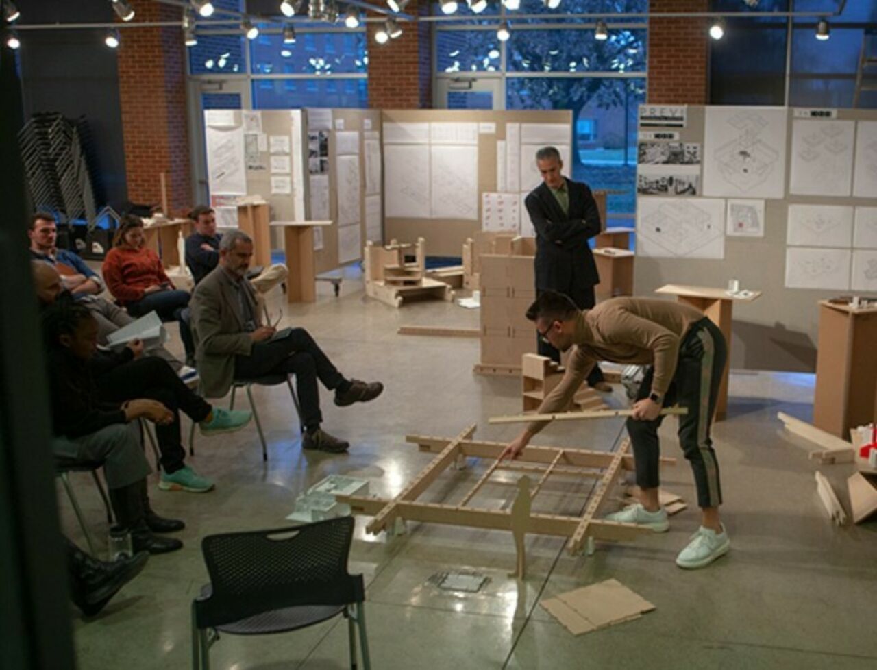 A male architecture student points to the base of his model on the floor as he explains the working foundation during reviews to the jury.