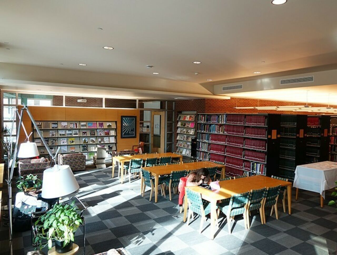 Inside view of the library with window light coming in from the left, students working at tables in the foreground, meeting space in the back, and stacks of books to the right.