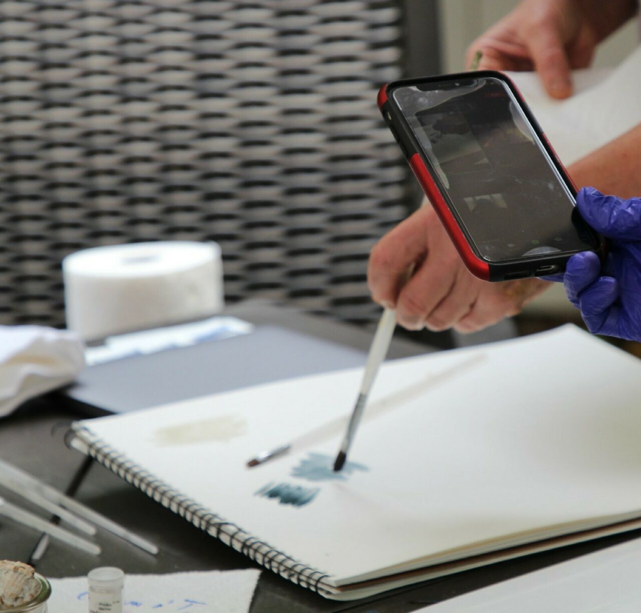 A gloved hand holding a smartphone to photograph paints being brushed onto paper at the Dye and Pigment Making Workshop
