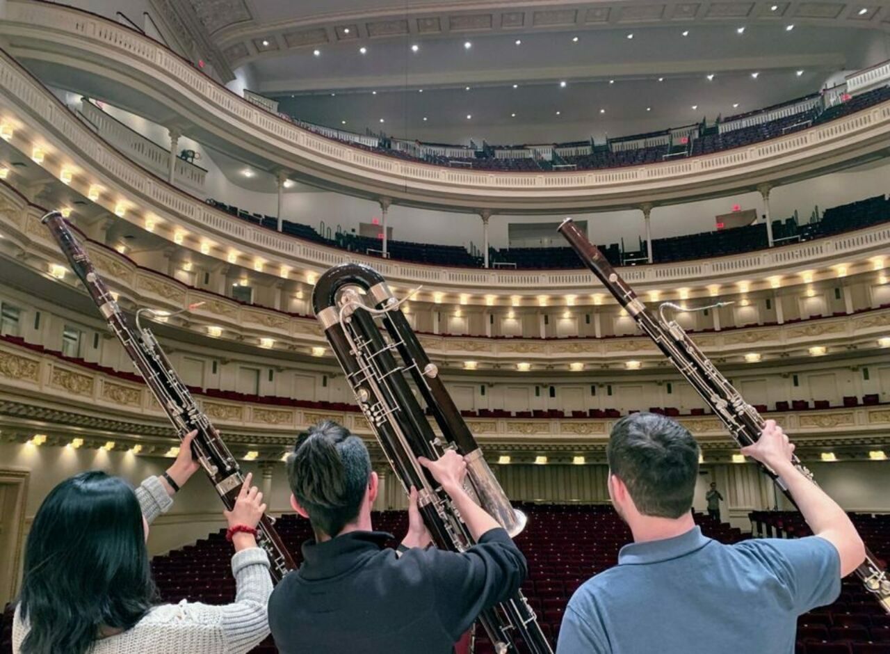 Three students hold up bassoons toward audience in auditorium, with backs to camera.