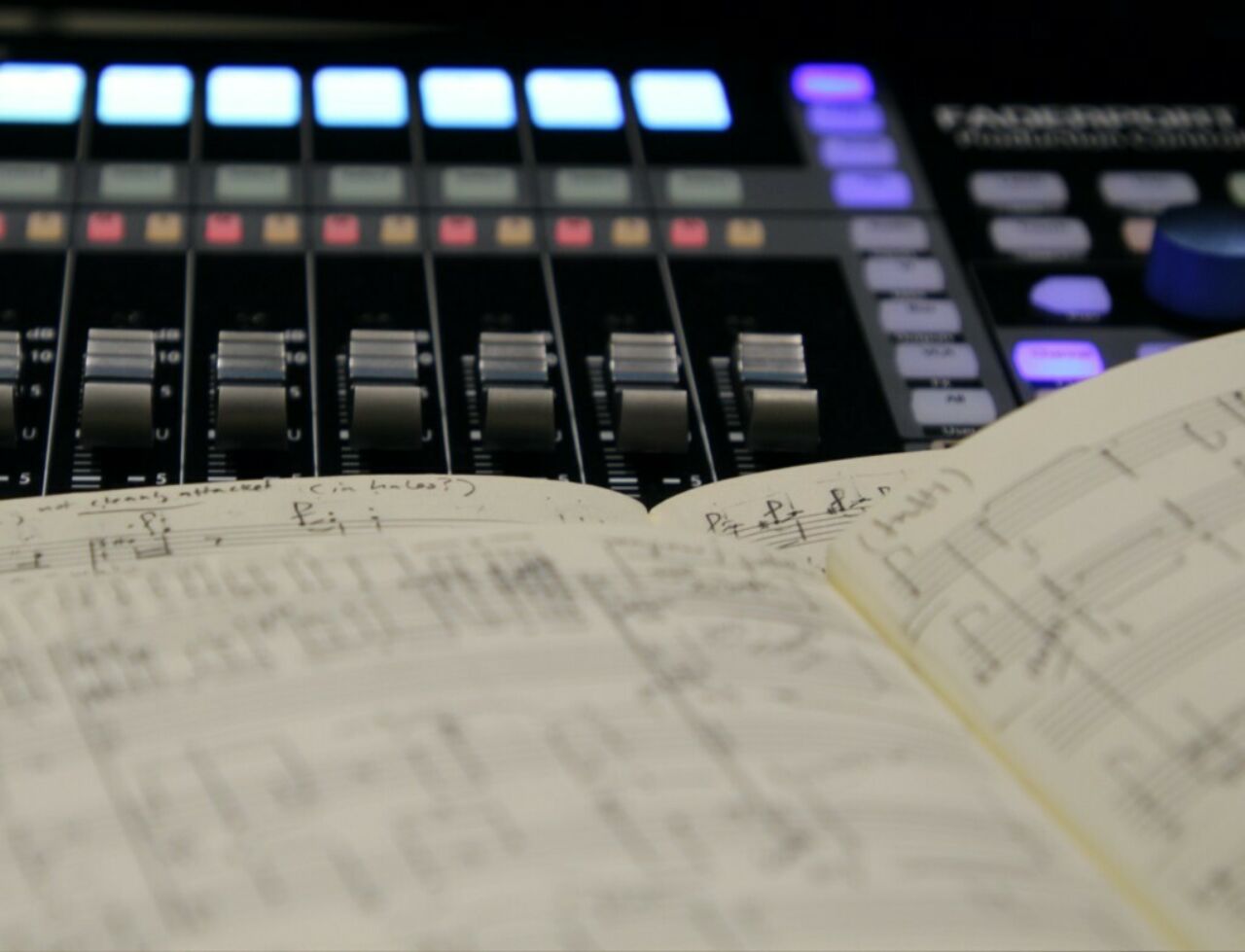 Close up image with blurred and sharp focus areas showing two hand-written music scores with a digital audio mixing board in the background.