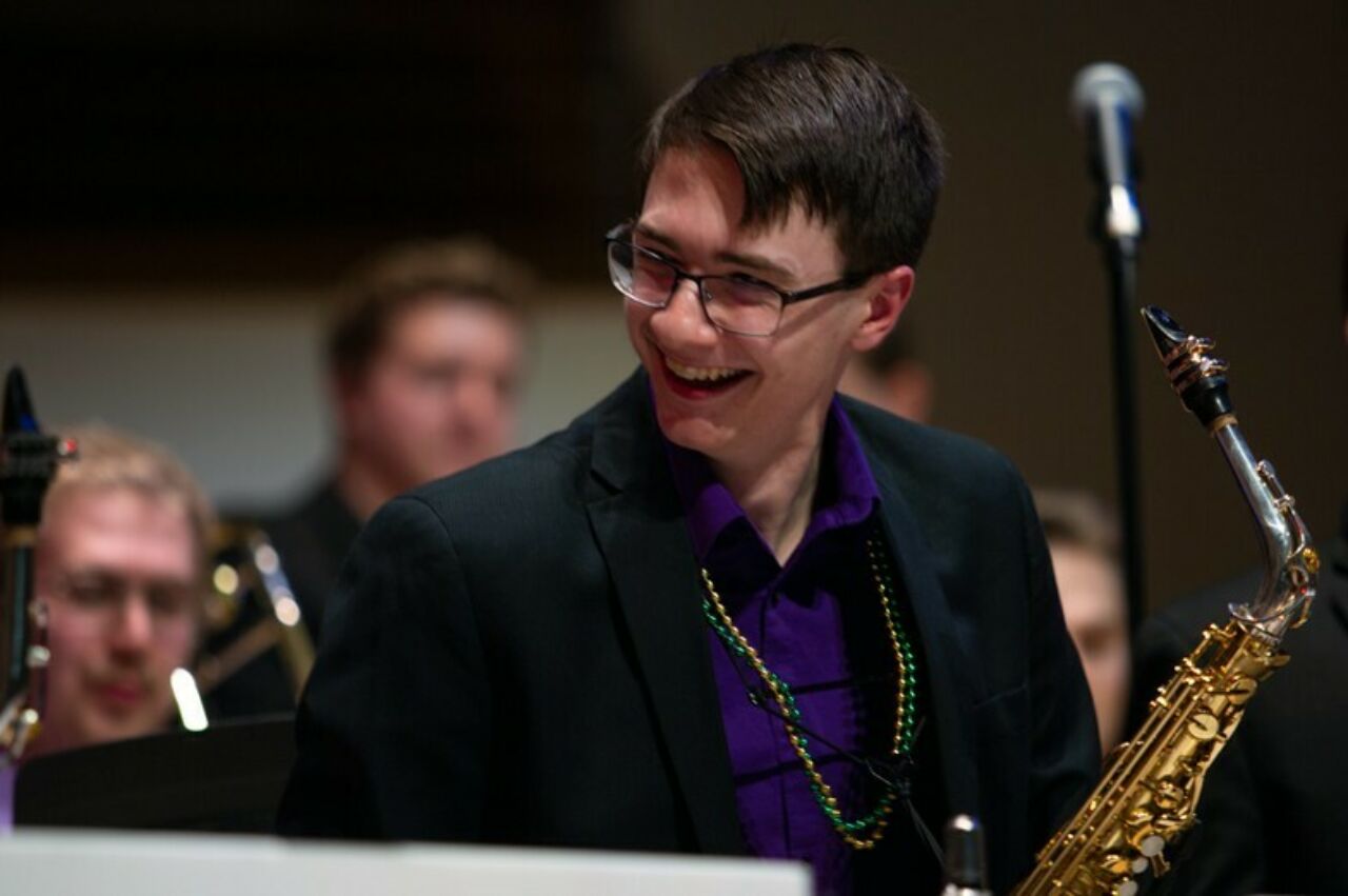 A jazz performer is holding is saxophone and his smiling face towards to another performer who is partially outside the photo