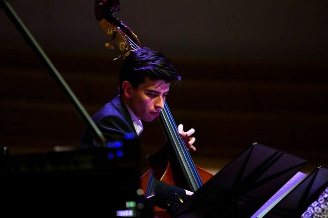 Bassist, seated behind piano and music stands playing.