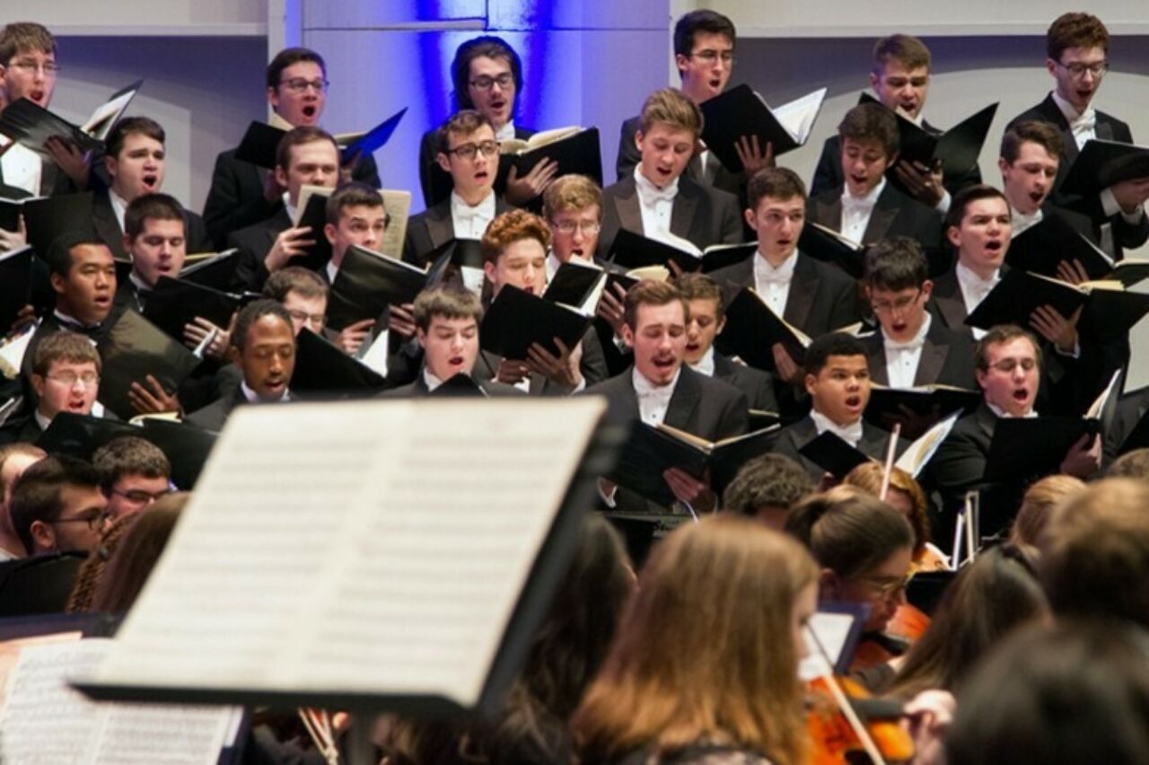 A blurred music stand occupies the foreground, in front of a large, formal student choir singing.