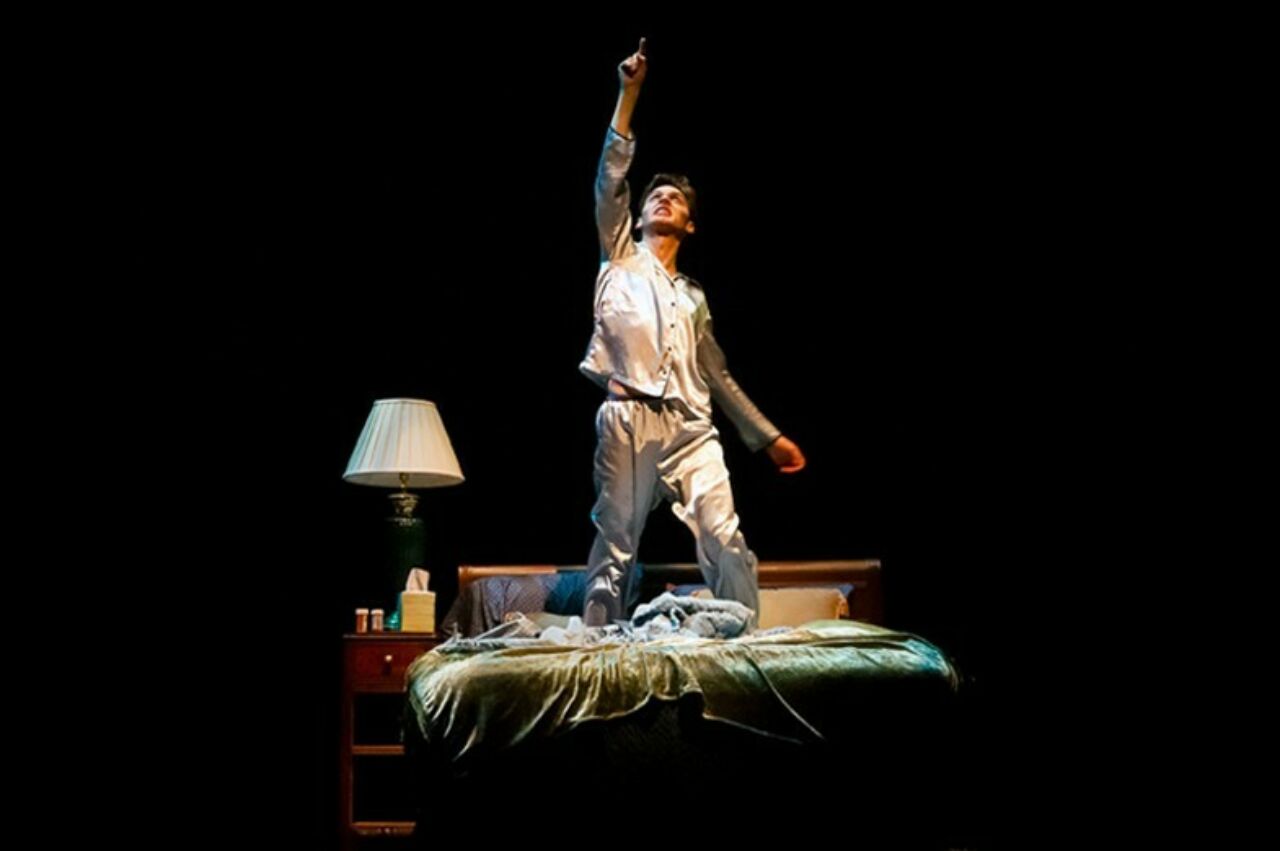 Student theatre performer kneeling on bed on a darkened stage with with arm outstretched overhead.