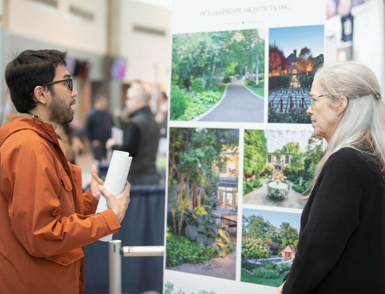 A student at left looks up while speaking to a woman at right in front of a landscape architecture firm's poster.