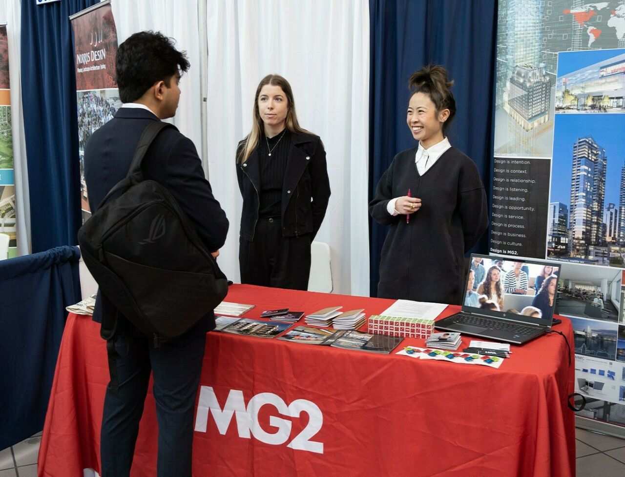 Two women stand behind a table with a red MG2 tablecloth on it while talking to a male student in front of them.