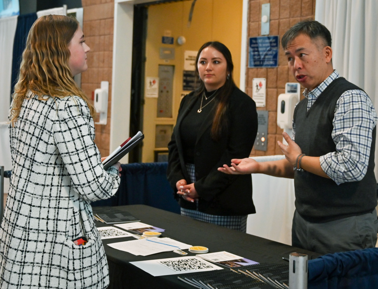 A Stuckeman School student at left holding a portfolio talks with two professional firm representatives who are standing behind a table at the 2024 Career Day in the Bryce Jordan Center.