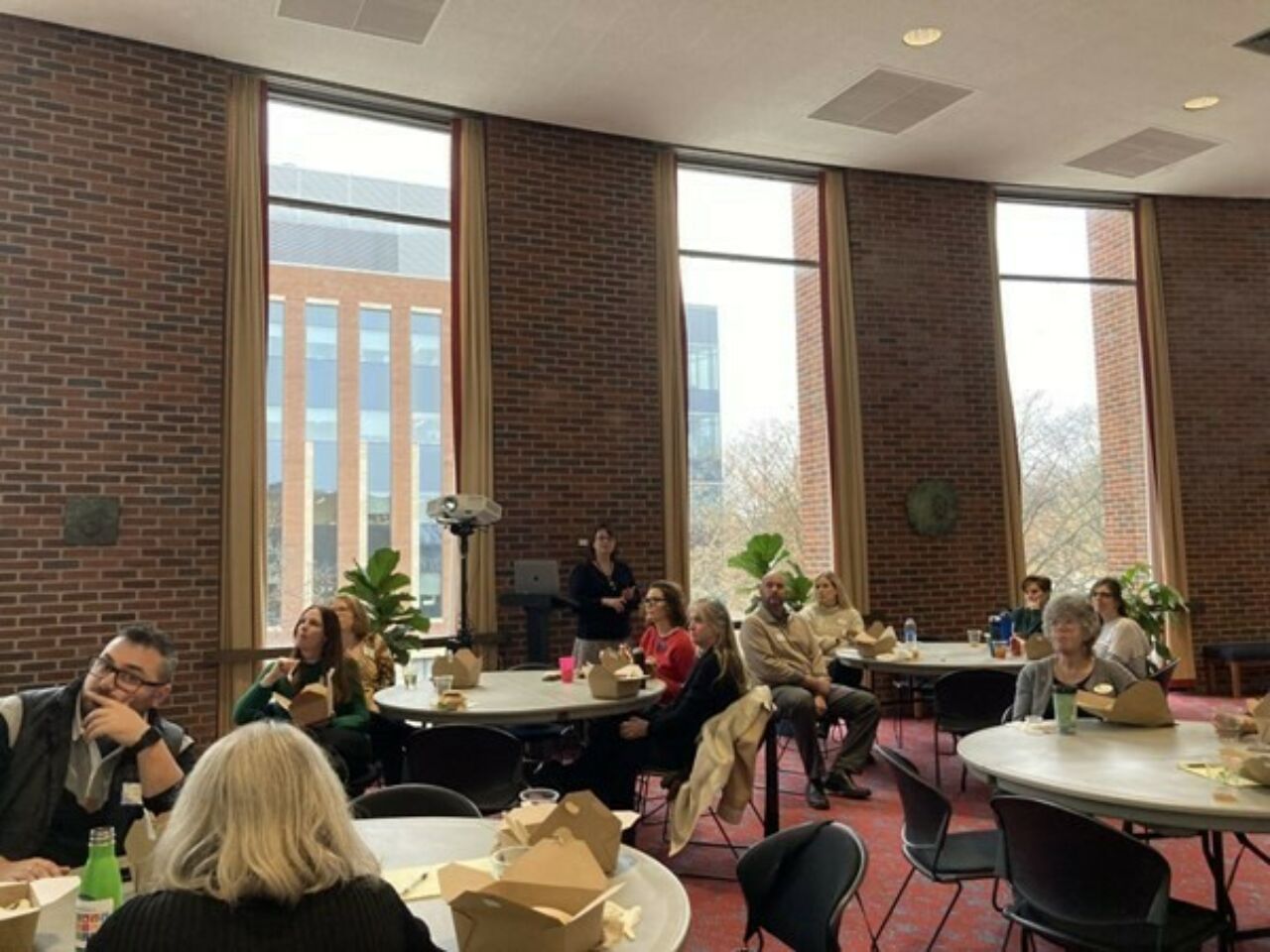 people seated a circular tables with box lunches in a room with brick walls, floor-to-ceiling windows, and red carpet.