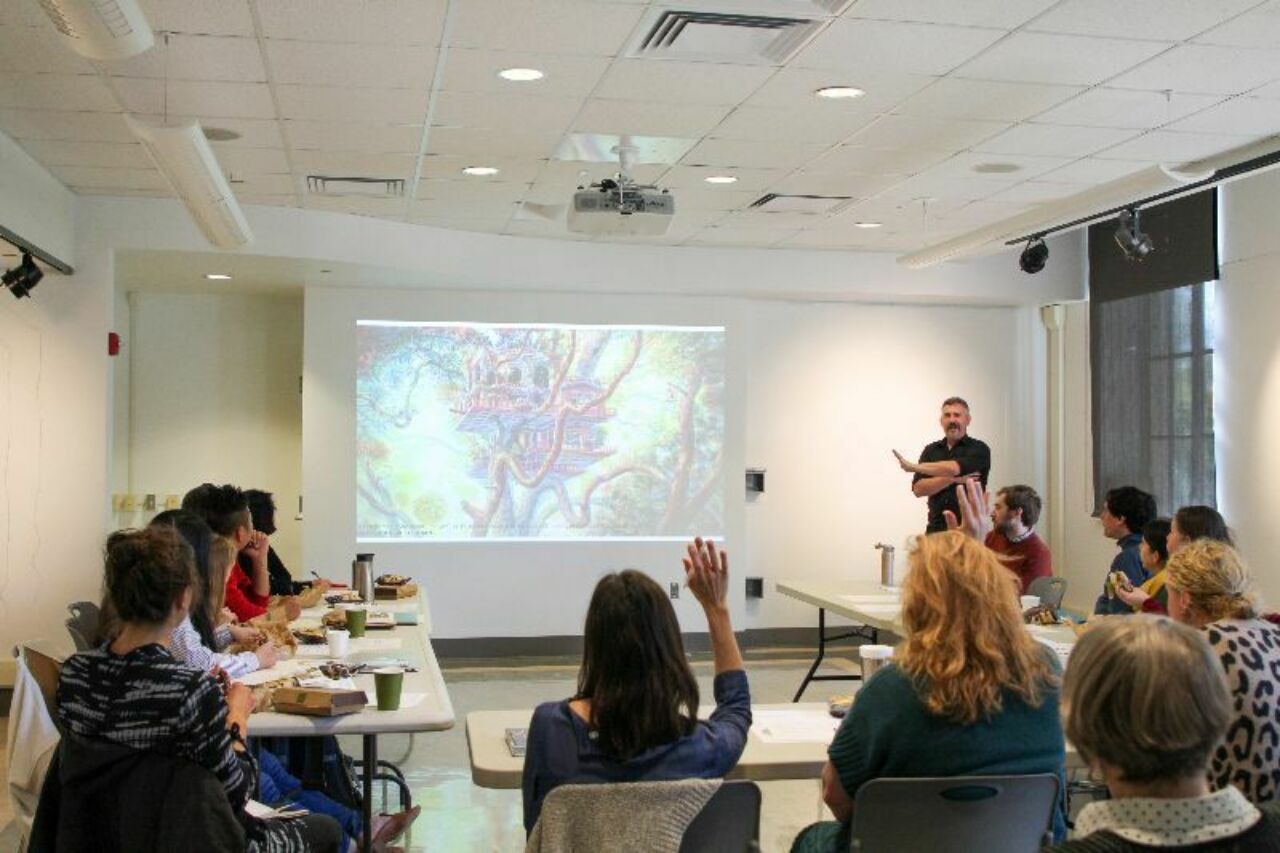Man giving a lecture in front of a classroom of people; one of the female attendees has her hand raised.