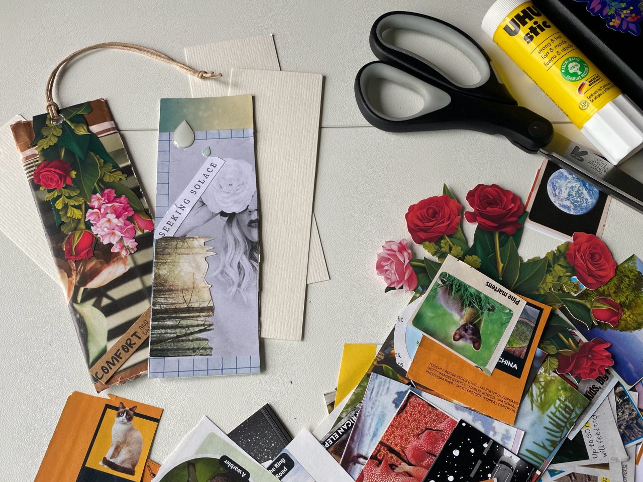 A photo showing several "collage" bookmarks on a table with scissors, glue stick, and pieces of art – the tools and materials to make the bookmarks.