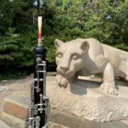 The half of oboe with the reed end is at the closer side of the image. The background is blurred Nittany Lion statue.