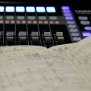 Close up image with blurred and sharp focus areas showing two hand-written music scores with a digital audio mixing board in the background.