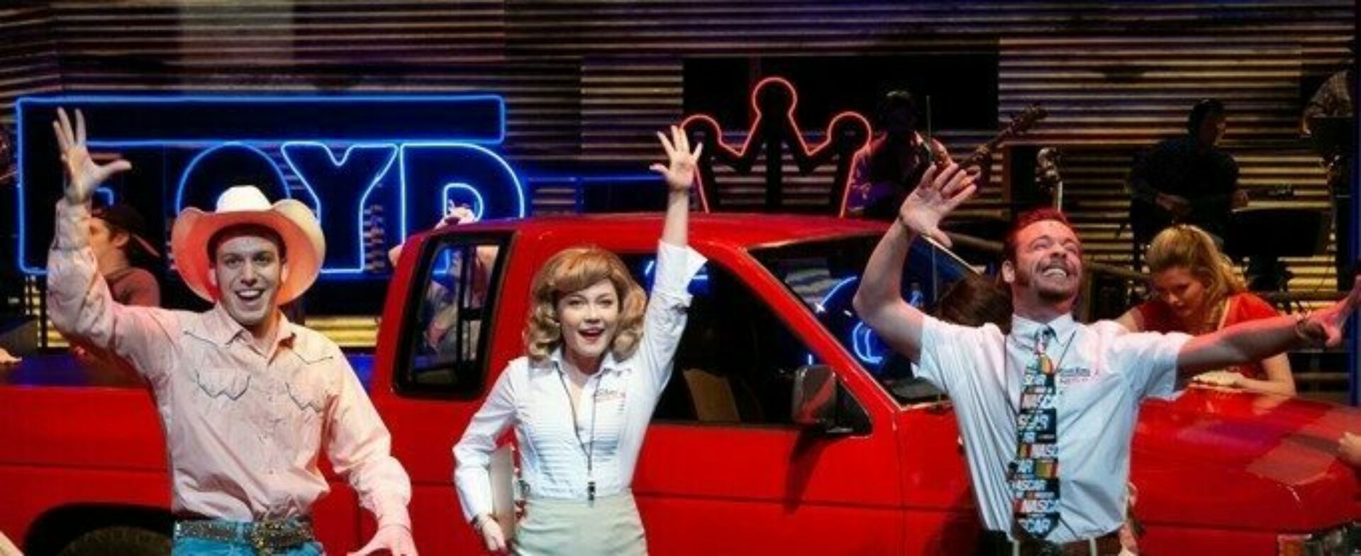 Theatre students dancing and singing in front of a bright red truck with neon lights in the background.