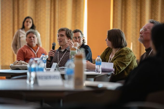 A group of people seated at tables with one man holding a microphone to his mouth