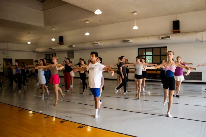 Group of theatre camp dancers in a studio facing a mirror with their arms held gracefully out to their sides and their right foot pointed behind them.