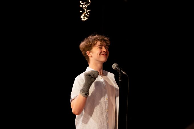 Individual theatre camp male smiling and standing on stage with a microphone in front of him and a sock on his one hand.