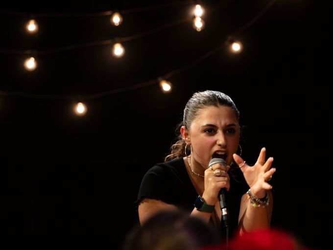 Individual theatre camp female singling into a microphone onstage bending low with one hand close to her serious face.