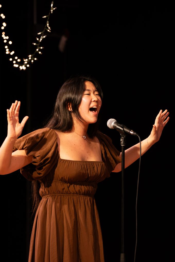 Individual theatre camp female singing into a microphone on stage with both arms up and open.