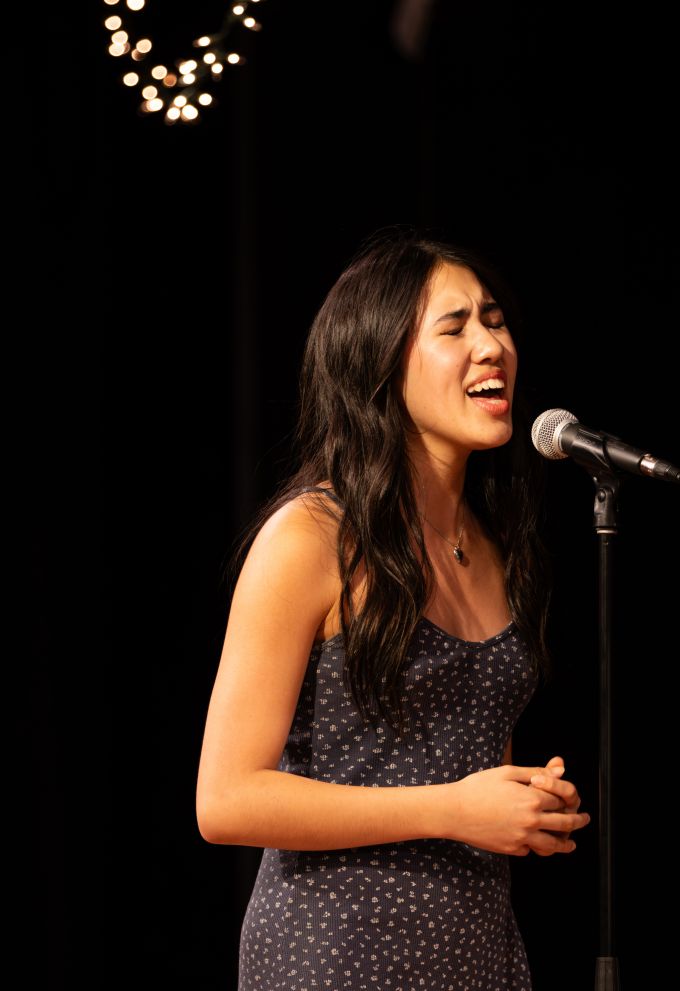 Individual female theatre camp student singing into a microphone with her eyes closed and arms around her waist.