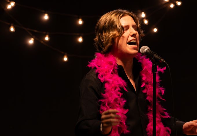 Individual male theatre camp student wearing a pink feather boa singing into a microphone.