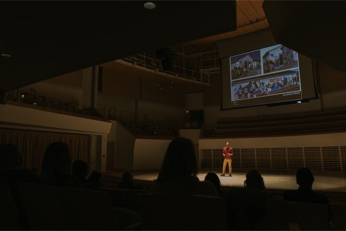 A darkly vignetted shot over the heads of an audience listening to a spotlit man on a large stage, with a presentation projected above and behind the stage level.
