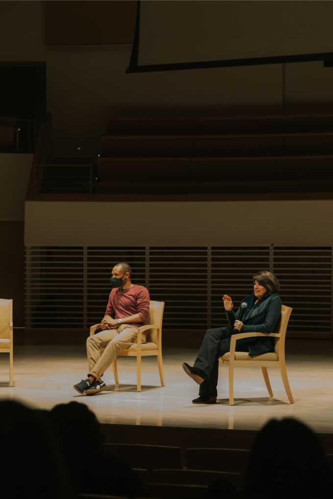 A man and woman sitting casually in chairs on a spotlit stage; the woman is holding a microphone and both people are wearing (COVID) safety masks.