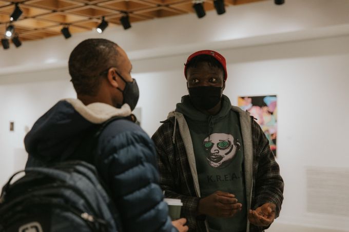 Closeup of a black student artist, wearing a sweatshirt with a caricature illustrated head on the chest, facing the camera and speaking to another black man wearing a blue, hooded puffy jacket.