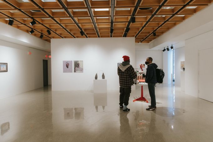 Two black men standing, turned away, in the white-walled, lattice-ceilinged, Zoller Gallery at Penn State's University Park campus. The gallery is very minimalistic, displaying just a few paintings on the white walls and sculptures on white plinths.